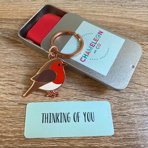 May include: A rose gold robin keyring with a red breast, brown back, and white belly. The keyring is in a silver tin with a blue label that says "Chameleon and Co.". A blue card with the text "Thinking of You" is in the foreground.