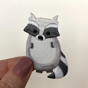 May include: A grey and black embroidered iron-on patch of a raccoon with a black nose and eyes.