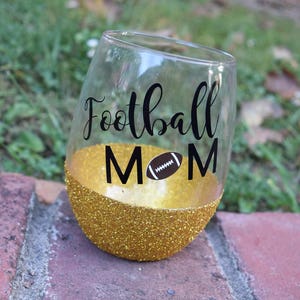 May include: A clear glass wine glass with a gold glitter rim and the words "Football Mom" with a football icon in black lettering.