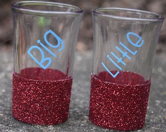 May include: Two clear shot glasses with red glitter around the bottom. The glass on the left says "Big" in blue lettering and the glass on the right says "Little" in blue lettering.
