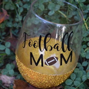 May include: A gold glitter wine glass with a black vinyl decal that says "Football Mom" with a brown football graphic.