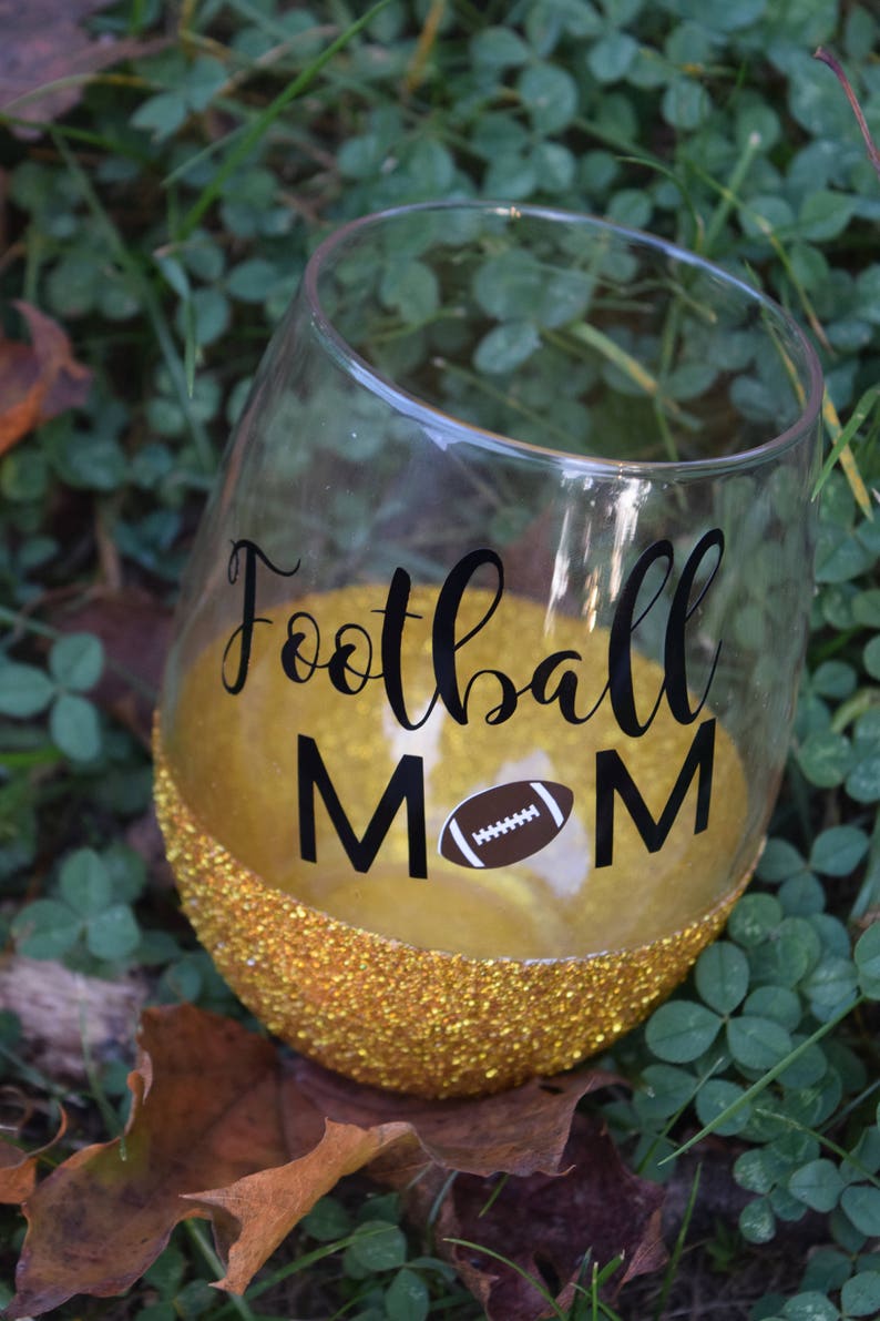 May include: A gold glitter wine glass with a black vinyl decal that says "Football Mom" with a brown football graphic.