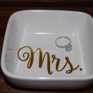 Puede incluir: Plato de cer&aacute;mica blanco para anillos con la palabra "Mrs." escrita en letras doradas. Un anillo de compromiso de diamantes est&aacute; sentado en el plato.