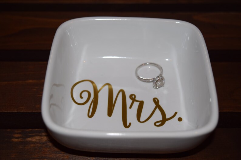 Puede incluir: Plato de cer&aacute;mica blanco para anillos con la palabra "Mrs." escrita en letras doradas. Un anillo de compromiso de diamantes est&aacute; sentado en el plato.