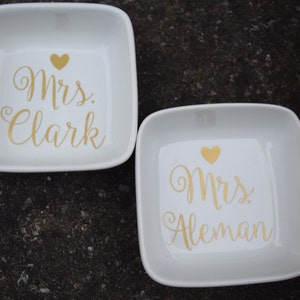 Puede incluir: Dos platos de anillos de cer&aacute;mica blanca con letras doradas. El primer plato dice "Mrs. Clark" y el segundo plato dice "Mrs. Aleman".