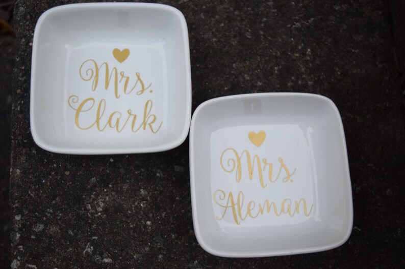 Puede incluir: Dos platos de anillos de cer&aacute;mica blanca con letras doradas. El primer plato dice "Mrs. Clark" y el segundo plato dice "Mrs. Aleman".