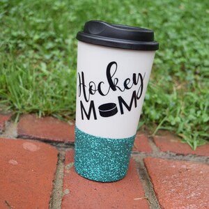 May include: White travel mug with a black lid and a teal glitter bottom. The mug has black lettering that says "Hockey Mom" with a black hockey puck graphic.