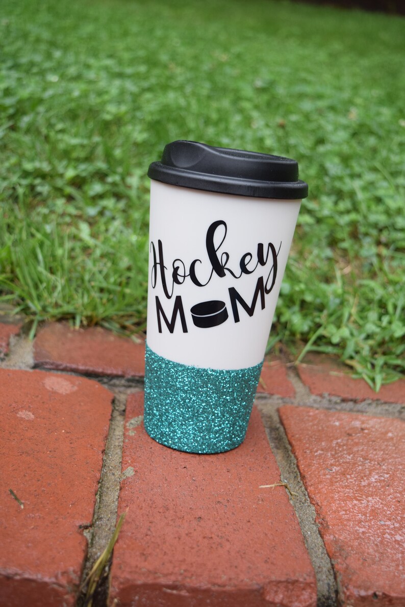 May include: White travel mug with a black lid and a teal glitter bottom. The mug has black lettering that says "Hockey Mom" with a black hockey puck graphic.
