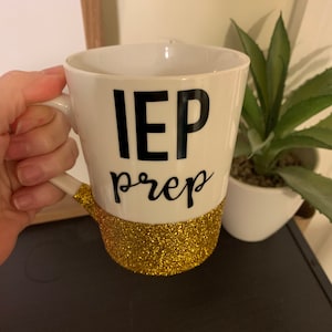 Pu&ograve; includere: Una tazza in ceramica bianca con la scritta "IEP prep" in nero e una fascia di glitter dorati intorno alla base.