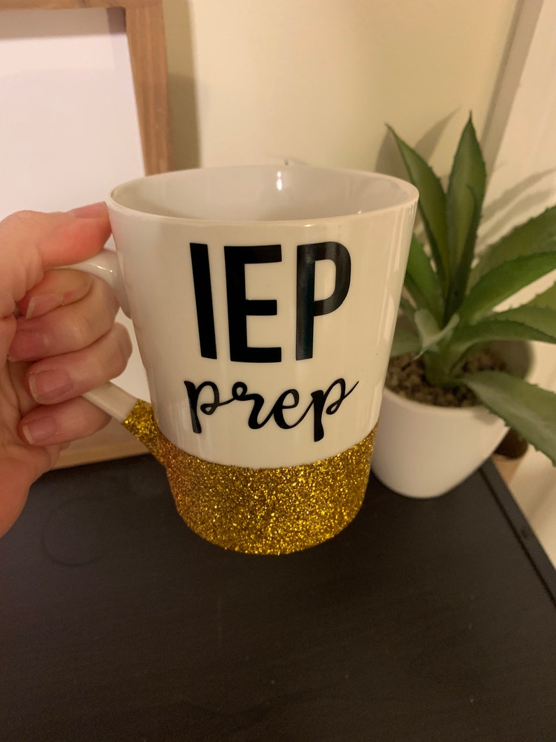 Pu&ograve; includere: Una tazza in ceramica bianca con la scritta "IEP prep" in nero e una fascia di glitter dorati intorno alla base.