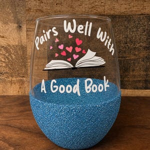 May include: A clear stemless wine glass featuring a blue glitter base and white text that reads Pairs Well With A Good Book. Above the text is an illustration of an open book with small pink and red hearts floating upward from the pages.