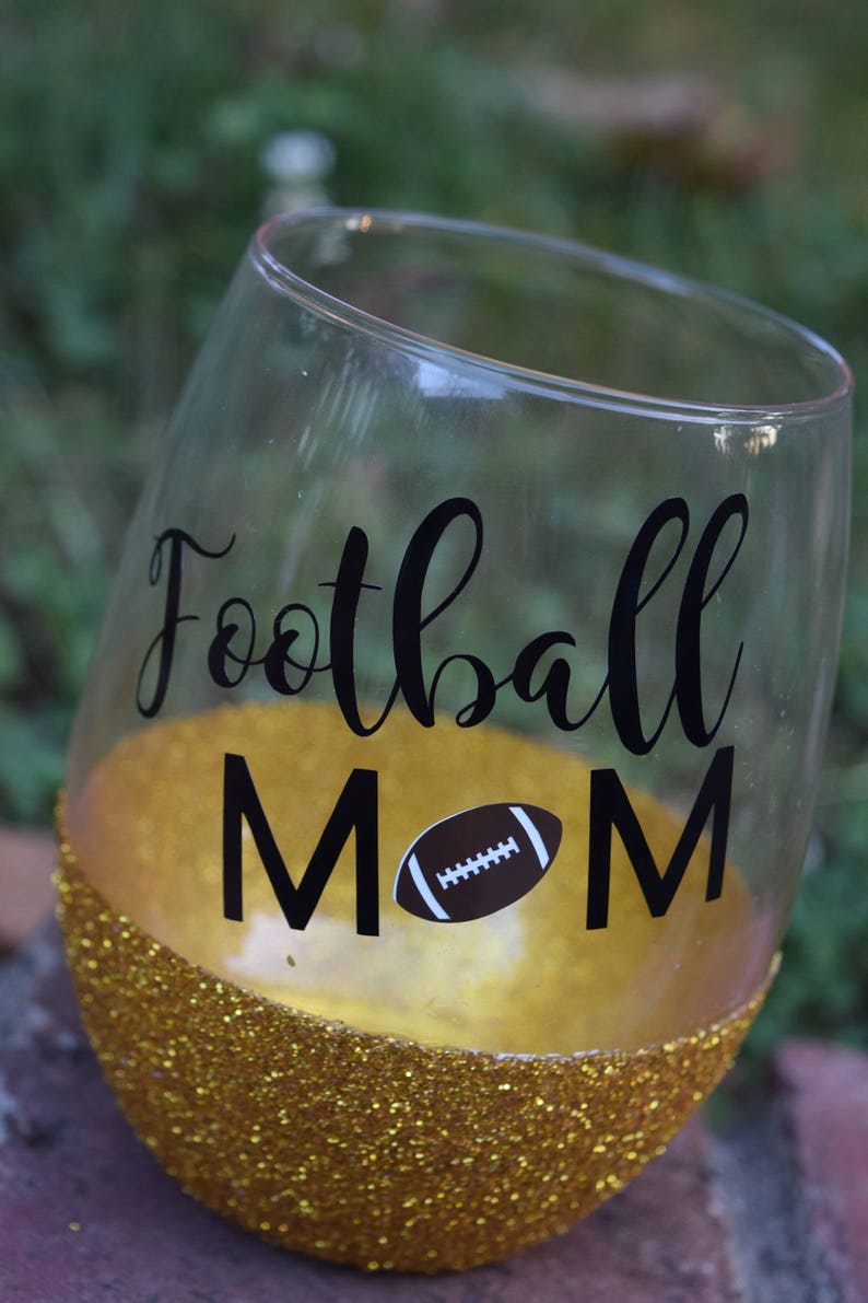 May include: A gold glitter wine glass with a black vinyl decal that says "Football Mom" with a football graphic.