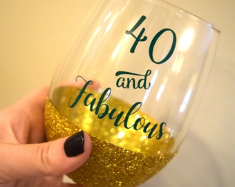 Può includere: Un bicchiere da vino in vetro trasparente con un fondo di glitter dorati. Il bicchiere ha un adesivo in vinile verde che dice "40 e favoloso".