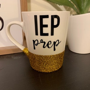 Pu&ograve; includere: Una tazza da caff&egrave; bianca con la scritta "IEP prep" in nero e una fascia di glitter dorati intorno alla base.