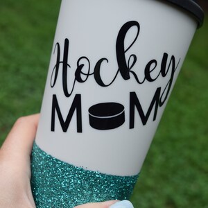 May include: A white travel mug with a black lid and a teal glitter bottom. The mug has the words "Hockey Mom" written in black cursive with a black hockey puck graphic.