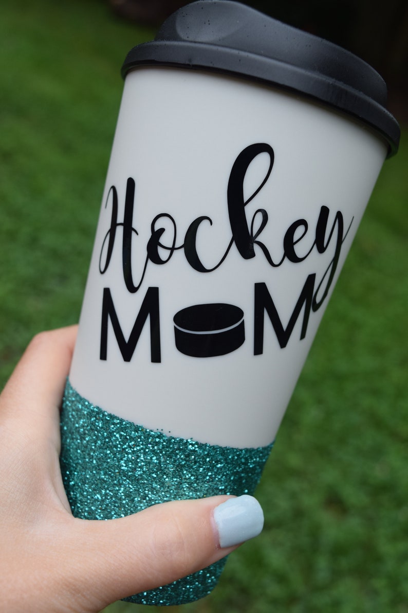 May include: A white travel mug with a black lid and a teal glitter bottom. The mug has the words "Hockey Mom" written in black cursive with a black hockey puck graphic.