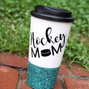May include: White travel mug with a black lid and a teal glitter bottom. The mug has black text that says "Hockey Mom" with a black hockey puck graphic.