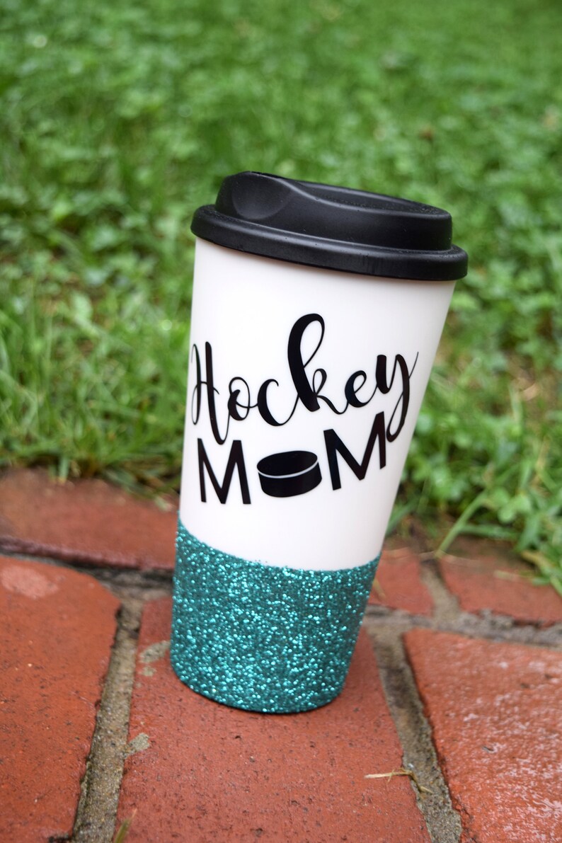 May include: White travel mug with a black lid and a teal glitter bottom. The mug has black text that says "Hockey Mom" with a black hockey puck graphic.