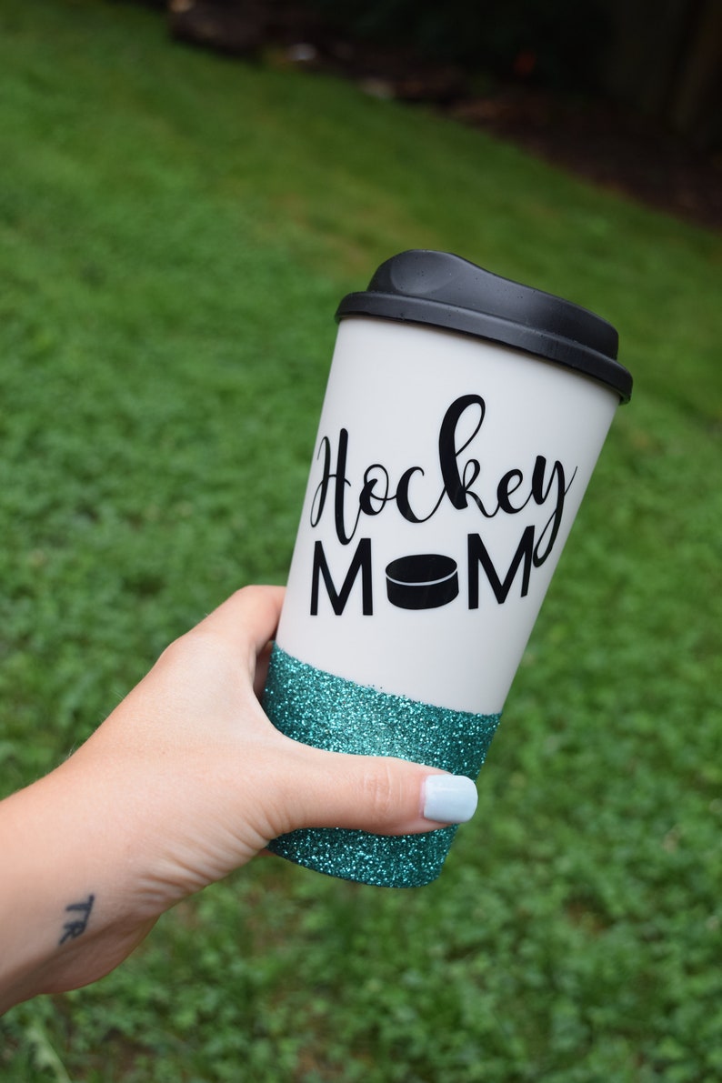 May include: White travel mug with a black lid and a teal glitter band. The mug has the text "Hockey Mom" with a black hockey puck graphic.