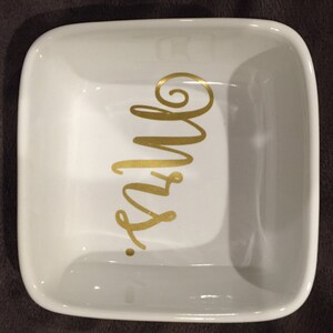 Puede incluir: Plato cuadrado de cer&aacute;mica blanca con la inscripci&oacute;n 'Mrs.' en dorado. El plato es cuadrado y tiene un borde ligeramente elevado.