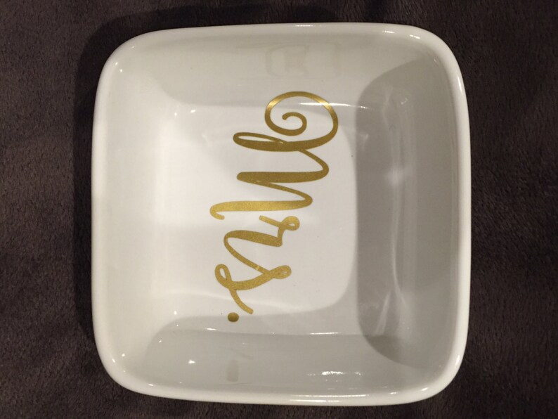 Puede incluir: Plato cuadrado de cer&aacute;mica blanca con la inscripci&oacute;n 'Mrs.' en dorado. El plato es cuadrado y tiene un borde ligeramente elevado.