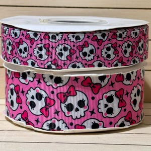 May include: A spool of pink grosgrain ribbon featuring a repeating pattern of white skulls with black eyes and pink bows. The ribbon has a textured, ribbed surface and is wound into a cylindrical shape, displaying the skull print across the entire width.