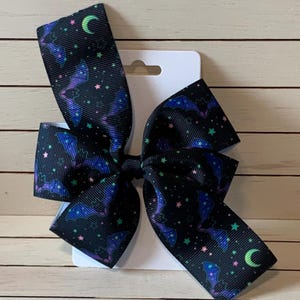 May include: A black hair bow with a purple, green, and pink bat and star pattern. The bow is attached to a white card.