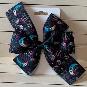 May include: A black hair bow with a colorful moon and mushroom print. The bow is tied in a large, symmetrical knot.