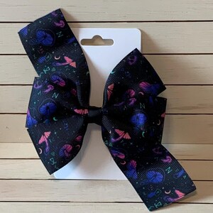 May include: A black hair bow with a pink, blue, and green print of moons, stars, and other witchy symbols. The bow is attached to a white card.