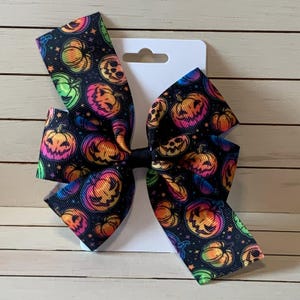 May include: A large Halloween hair bow featuring a repeating pattern of colorful jack-o'-lanterns on a black background. The bow is attached to a white card with a hang hole, perfect for display or gifting.