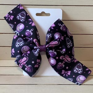 May include: A purple and black hair bow with a repeating pattern of purple hearts, moons, and eyes. The bow is attached to a white card with a hole for hanging.