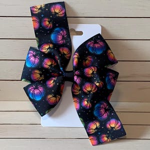 May include: A large hair bow featuring a black background with a pattern of colorful pumpkins. The pumpkins have a gradient of colors including blue, pink, orange, and yellow. The bow is attached to a white card.