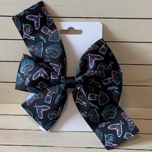 May include: A large black hair bow with a repeating pattern of colorful insects and other designs. The bow is attached to a white card with a hole for hanging. The bow is made of a ribbed fabric.