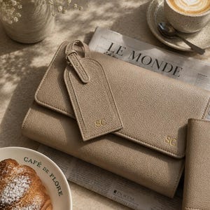 May include: A beige pebbled leather clutch with a matching luggage tag, both featuring the gold embossed initials SC. The clutch has a flap closure and is positioned on a newspaper next to a croissant, a cup of coffee, and tortoise shell sunglasses.