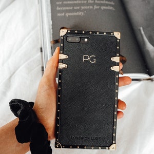 Puede incluir: Funda de teléfono de cuero negro con detalles dorados y las iniciales "PG" grabadas en la parte delantera. La funda tiene una inscripción "LOVER OF LUXE" en la parte inferior.