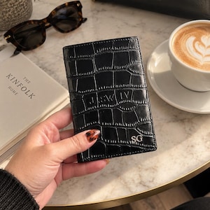 May include: A black passport holder with a glossy crocodile-embossed texture. The front features the embossed text "JASWLY" in the center and the initials "SG" in the bottom right corner. The rectangular accessory has visible stitching along the edges.