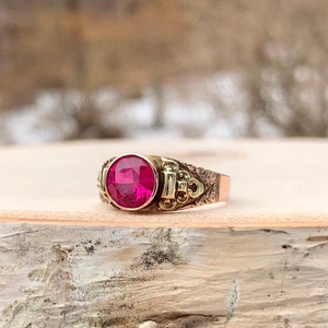 Puede incluir: Un anillo de oro con una gran piedra preciosa redonda de color rosa oscuro engastada en el centro. La banda est&aacute; decorada con intrincados dise&ntilde;os florales.