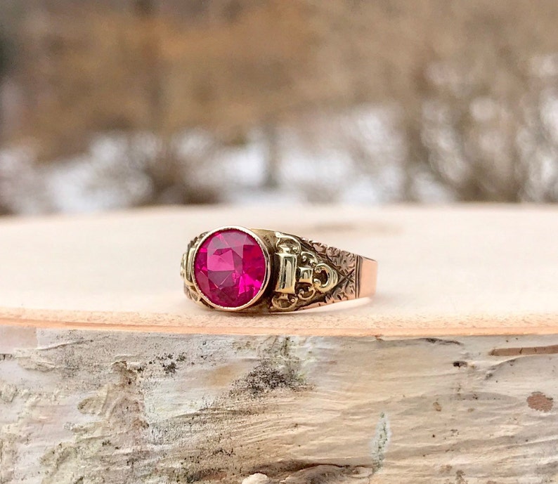 Puede incluir: Un anillo de oro con una gran piedra preciosa redonda de color rosa oscuro engastada en el centro. La banda est&aacute; decorada con intrincados dise&ntilde;os florales.