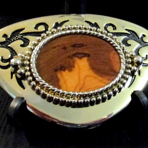 May include: A silver belt buckle with a decorative design featuring a circular inlay of wood with a brown and orange swirl pattern.