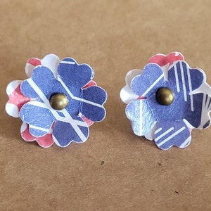 May include: A pair of blue and red paper flower earrings with gold accents. The flowers are layered and have a geometric pattern.