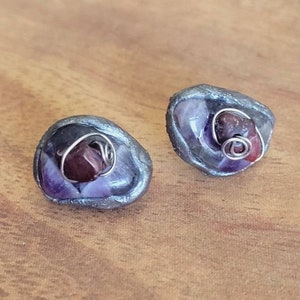 May include: A pair of earrings with a silver wire wrapped around a small red stone and set in a purple and gray stone. The earrings are shaped like a small, irregular oval.