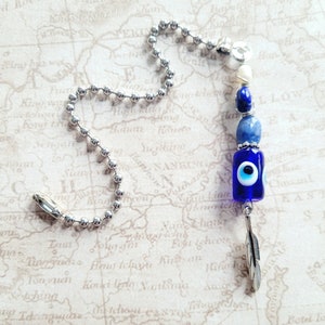 May include: A silver chain with a blue and white evil eye charm, a white shell bead, and a blue lapis lazuli bead. The charm is attached to a silver feather.