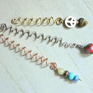 May include: Three spiral hair coils made of gold, silver, and copper-toned wire. The gold coil features a peace sign charm and a brown stone bead. The silver coil has a red patterned bead. The copper coil includes light green and blue beads.