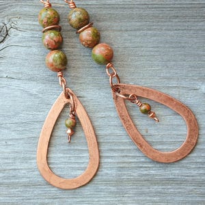 May include: Copper teardrop earrings with green and red stone beads. The earrings have a simple, open teardrop shape and are suspended from a chain with a cluster of round beads. The background is a weathered, gray wooden surface.