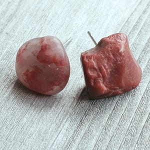 May include: A pair of stud earrings featuring natural stone designs. One earring is a rounded shape with red and pink hues, while the other is a rough, textured, reddish-brown stone. Both have silver posts.