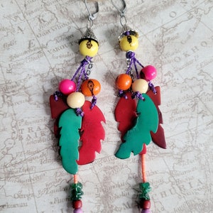 May include: A pair of dangle earrings with a colorful, layered design. The earrings feature green, red, and orange leaves, wooden beads, and a yellow bead at the top. The earrings are suspended from silver hooks.