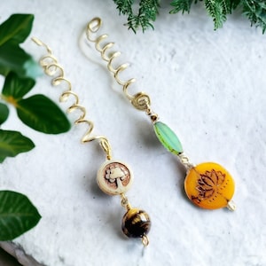 May include: Two gold hair coils with beads. One coil has a green glass bead and a round yellow bead with a lotus flower design. The other coil has a white bead with a tree design and a tiger's eye bead.