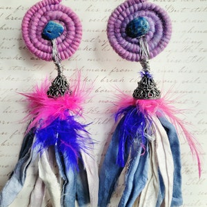 May include: A pair of handmade earrings with a bohemian style. Each earring features a spiral-shaped, purple fabric base with a blue stone center. Below, a silver filigree cap supports a tassel of blue, white, and denim fabric, accented with pink feathers.