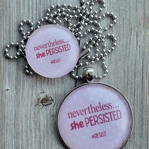 May include: A silver chain necklace with a pink glass pendant. The pendant has the text "nevertheless... she persisted #resist" printed on it.