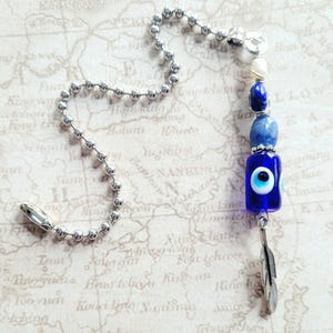May include: A silver-toned pull chain with a decorative charm. The charm features a blue glass bead with an evil eye design, blue and white beads, and a small silver feather pendant. The background is a map.